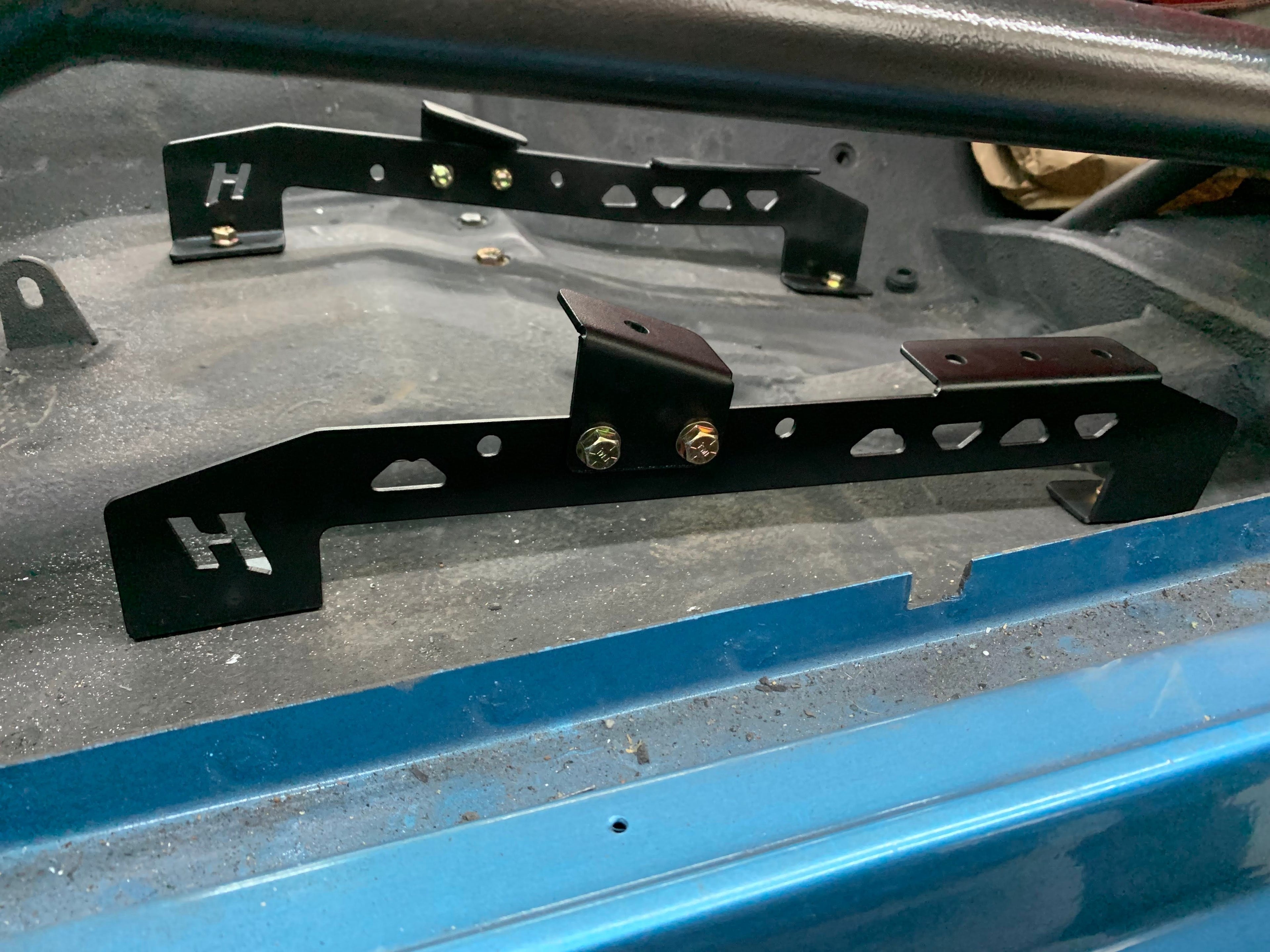 Henderson Race Shop black powder-coated steel seat mount brackets installed in a 2nd Gen Camaro or Firebird floor pan.