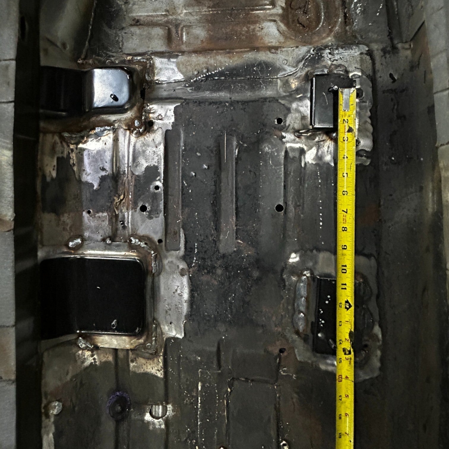 Henderson Race Shop Store seat mount brackets installed on a Chevy Nova floor pan with a measuring tape for spacing